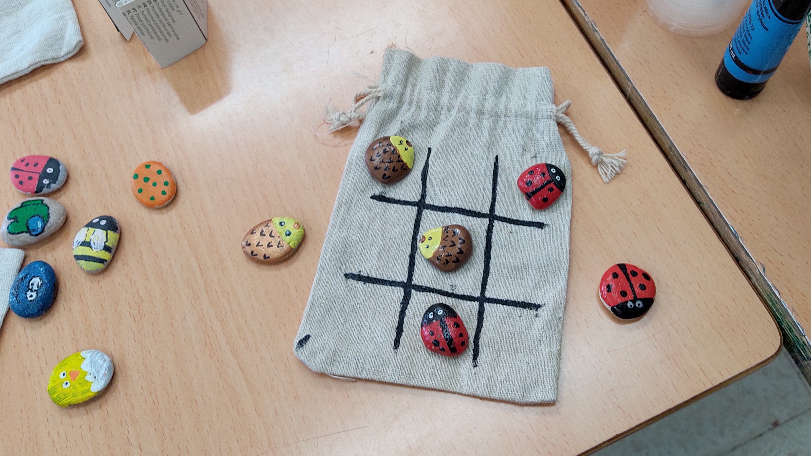 Tablero de Tres en Raya dibujado sobre madera, con piedras redondeadas pintadas con mariquitas y formas abstractas como fichas